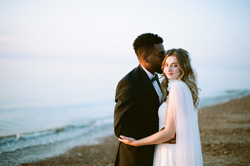 Groom kissing bride's cheek on beach during golden hour