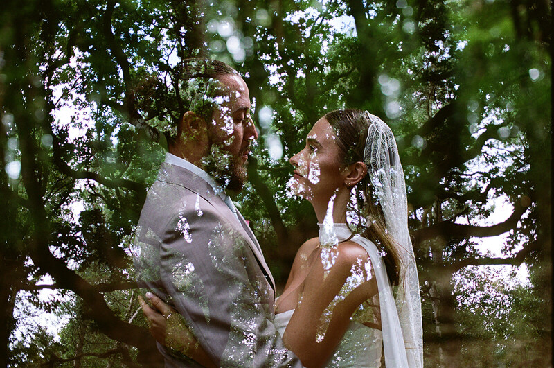 Bride and groom in wedding attire surrounded by tree canopy with soft, dappled natural light filtering through leaves