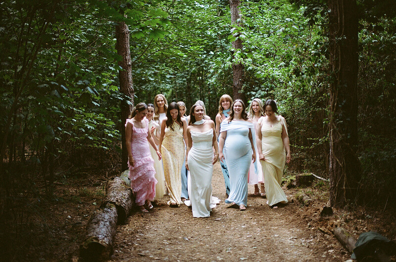 Group of women in pastel formal wear standing together on a forest path lined with tall trees