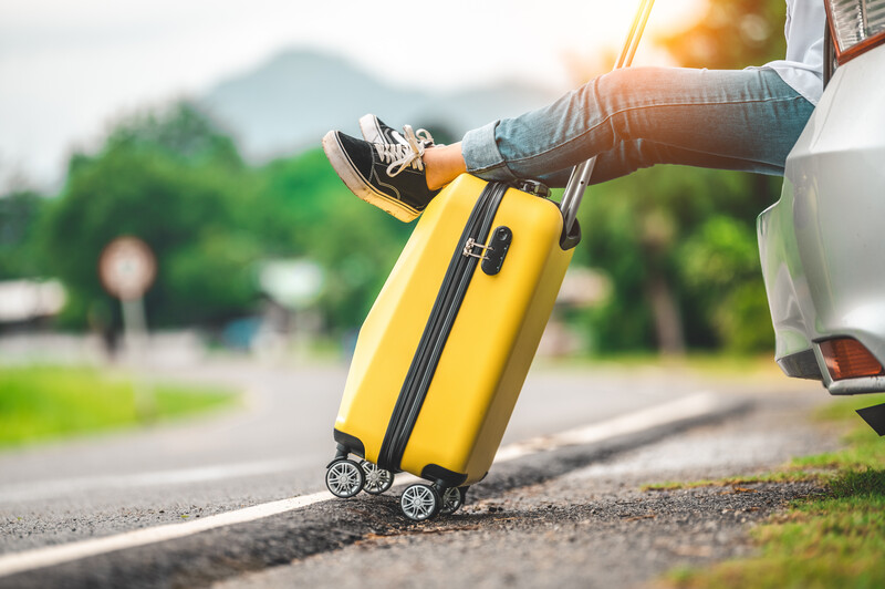 Person's legs dangling from vehicle window with yellow rolling suitcase below on road