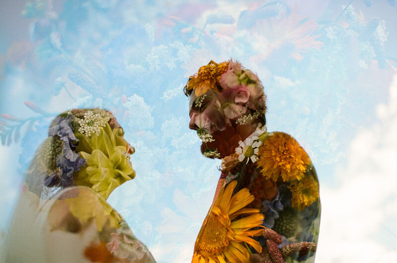 Double exposure portrait merging two faces with wildflower and daisy overlays against a soft sky