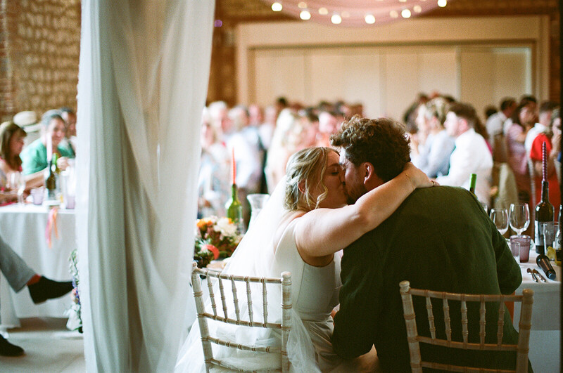 Bride and groom seated at wedding reception table, embracing while guests mingle in background