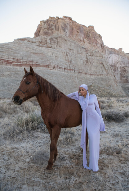 Woman in lavender dress standing beside brown horse against layered rock formation