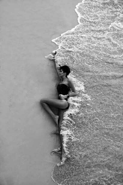 Black and white overhead shot of a shirtless man lying in shallow ocean waves on a beach