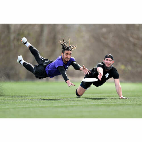 Female ultimate frisbee player diving horizontally to catch disc while defender crouches nearby on grass field