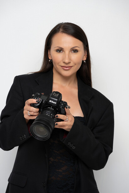 Professional photographer holding a DSLR camera, wearing black blazer against white backdrop
