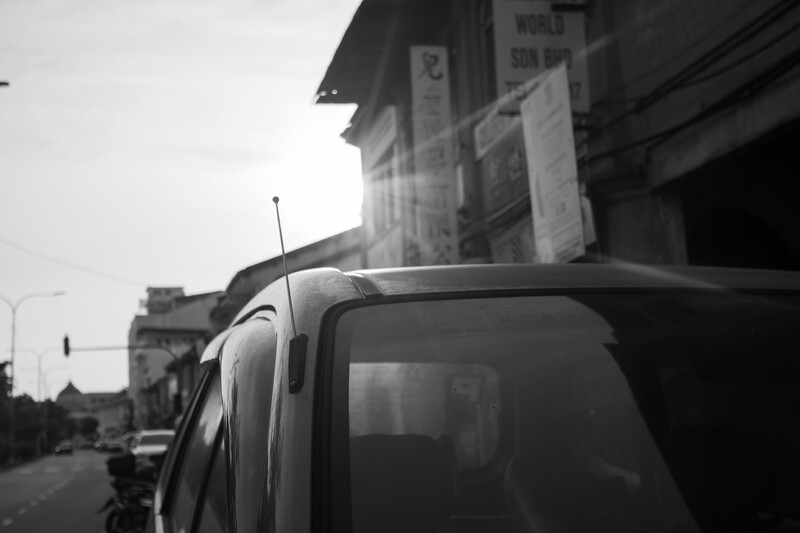 Black and white photograph of a vintage car's hood and windshield with urban street scene in background