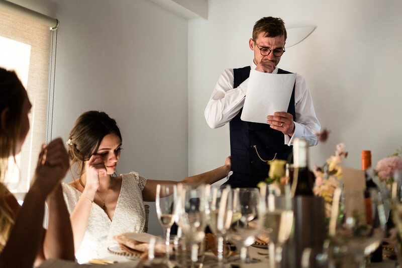 Man in vest and glasses reviews document while woman at desk listens during what appears to be a professional meeting or critique session