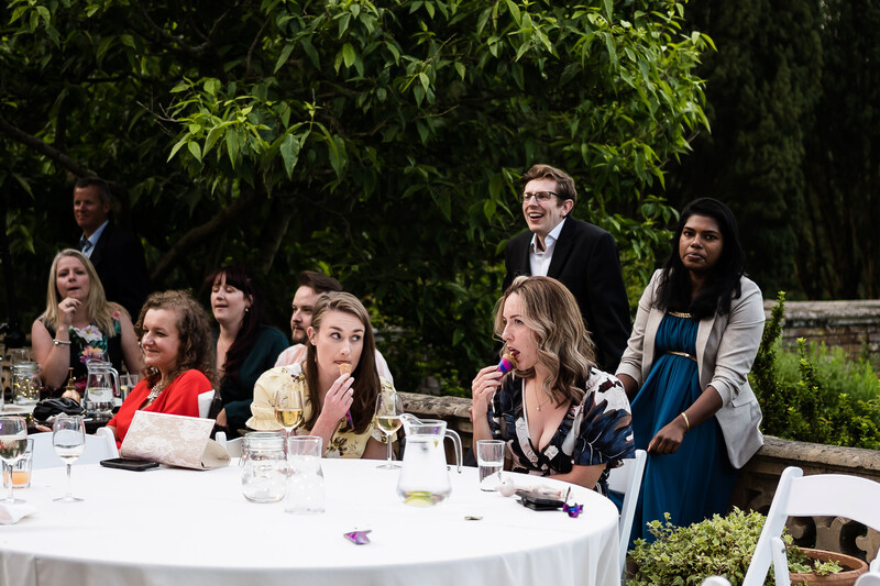 Group of people seated and standing around a white round table during an outdoor event with green foliage backdrop