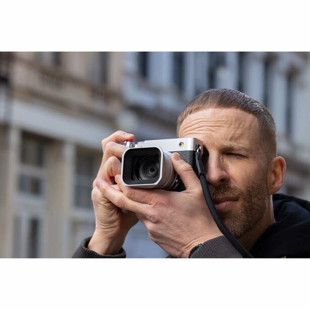 Bearded man holding compact camera to eye while composing shot, urban building backdrop