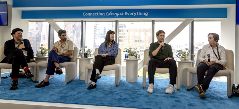 Five panelists seated in modern white chairs on blue carpet during a discussion panel with city skyline visible through windows behind them