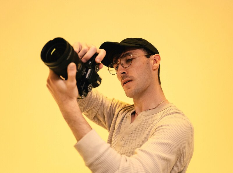 Photographer holding mirrorless camera to eye while wearing glasses against yellow background