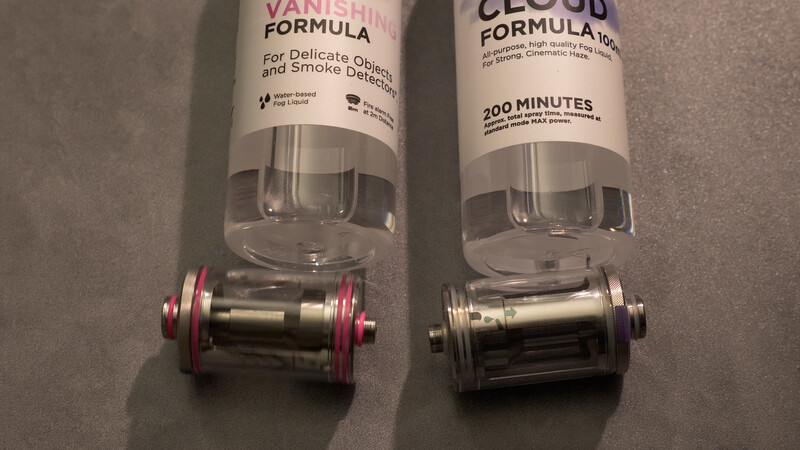 Two cylindrical vaping devices with clear tanks and colored accents placed side by side on a gray surface