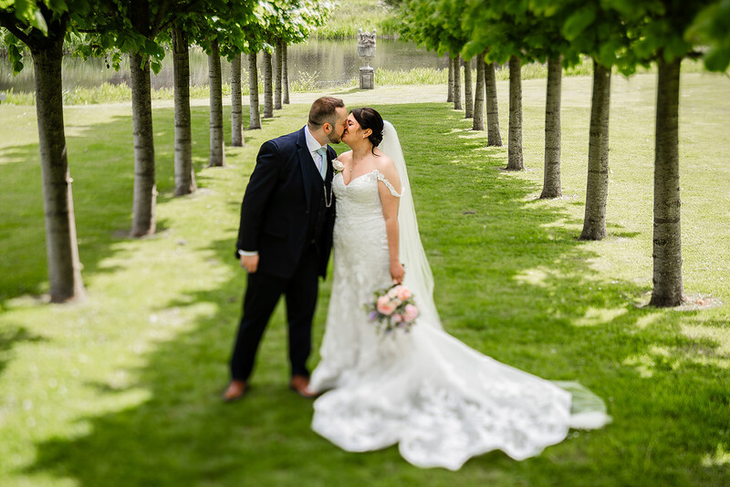 Bride and groom kissing on tree-lined grass pathway during outdoor wedding portrait session