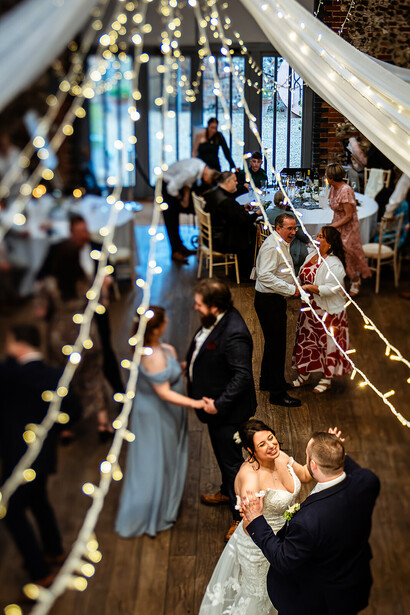 Wedding ceremony in a rustic venue with string lights creating a bokeh-filled foreground and bride and groom exchanging vows in sharp focus.