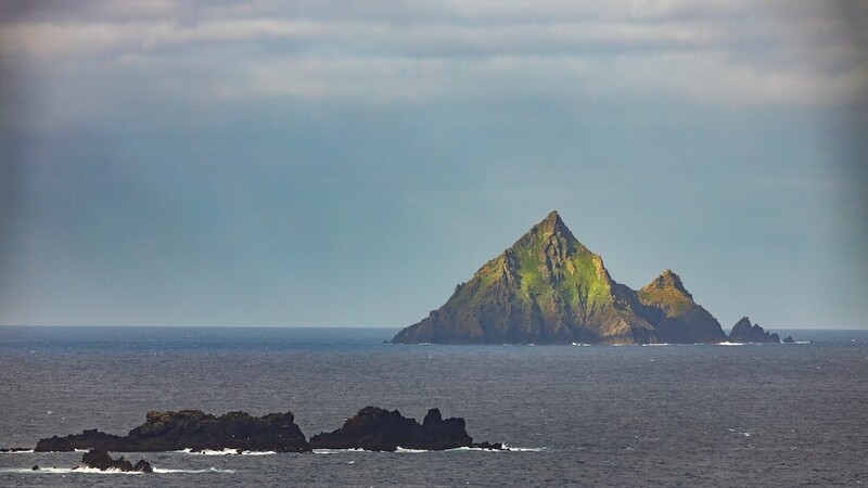 Rocky island with steep peaks rising from calm ocean waters under partly cloudy sky