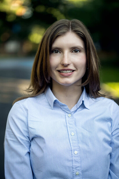 Woman with shoulder-length brown hair wearing light blue button-up shirt against blurred outdoor background