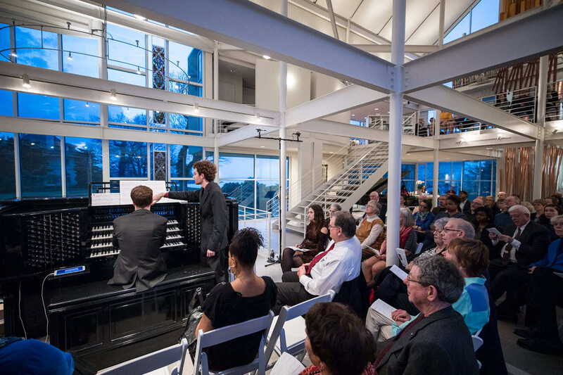 Pianist performing in a modern concert hall with audience members seated and standing throughout the multi-level space.