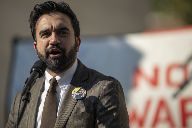 Man with beard speaking at microphone wearing gray suit jacket and pin