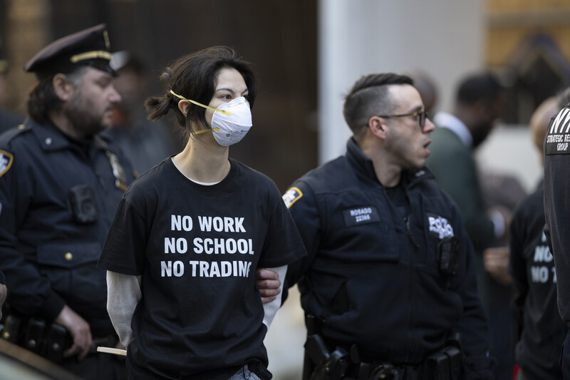 Woman wearing face mask and black shirt with white text being escorted by uniformed officers indoors