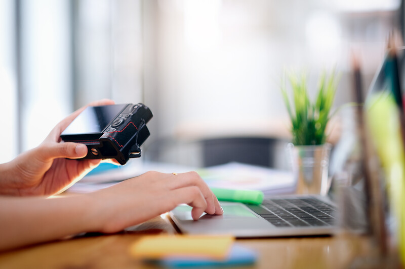 Photographer's hand holding a camera controller above a desk workspace with laptop and mouse