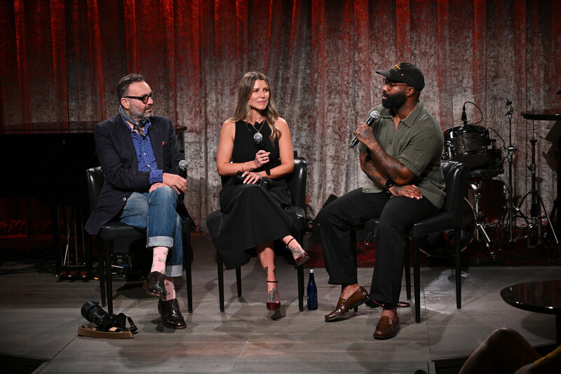 Three panelists seated on stage during a discussion, with professional lighting and backdrop