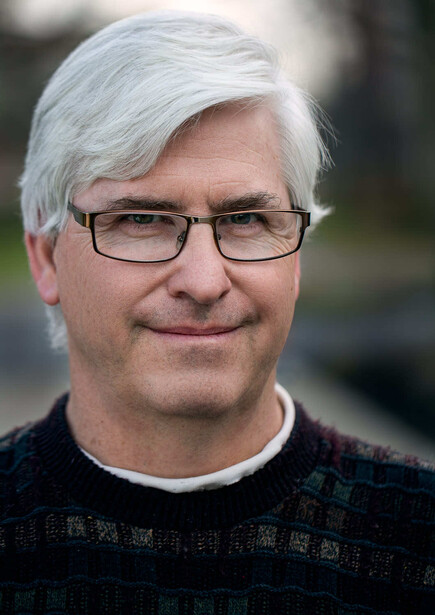 Headshot portrait of a man with white hair and glasses wearing a dark textured shirt