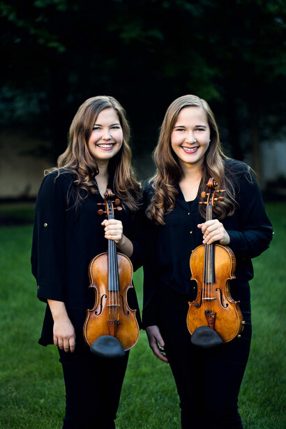 Two young women holding violins, posed outdoors against a blurred green background