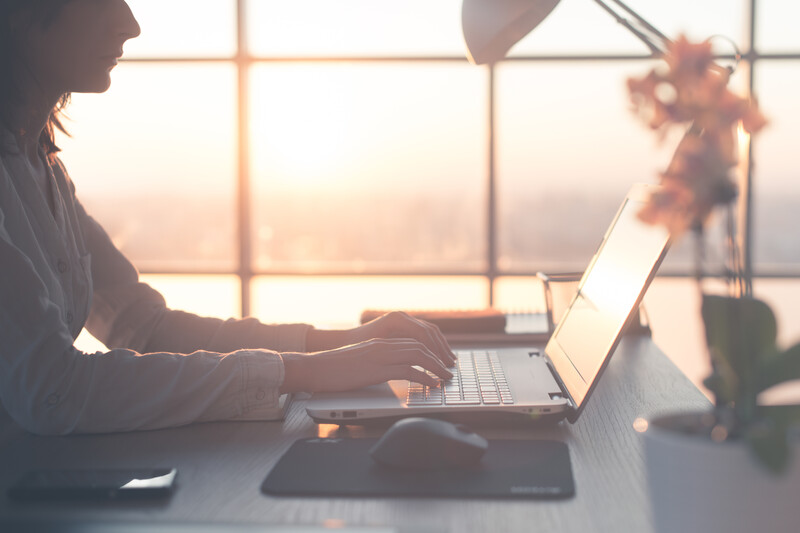 Woman working at laptop by large window during golden hour sunrise or sunset
