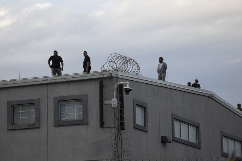 Multiple people standing on flat roof of gray industrial building with barbed wire coiled on top