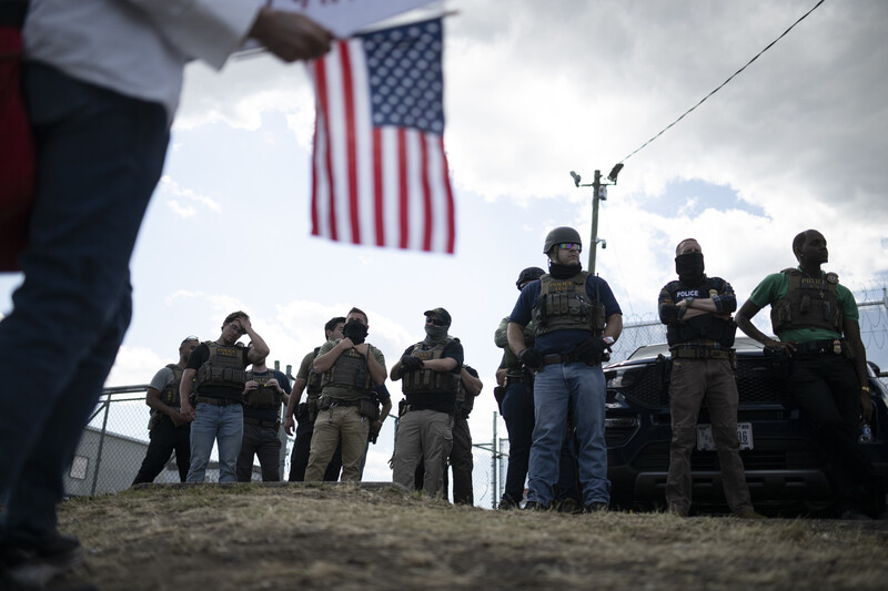 Armed personnel standing in formation beneath an American flag against overcast sky