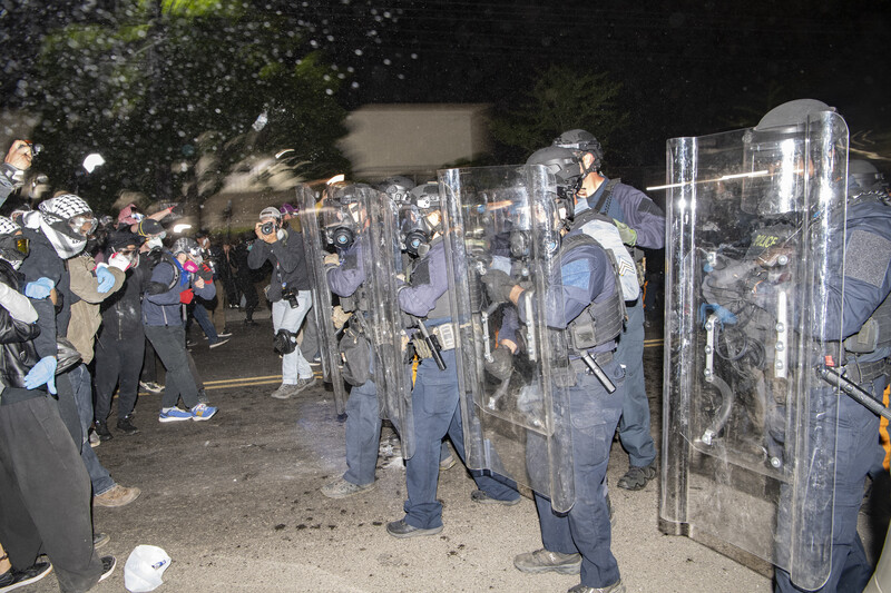 Police in riot gear facing crowd at nighttime street protest with confetti in air