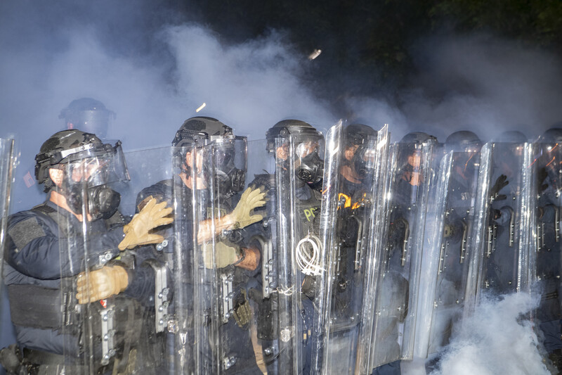 Riot police in protective gear holding shields amid tear gas clouds