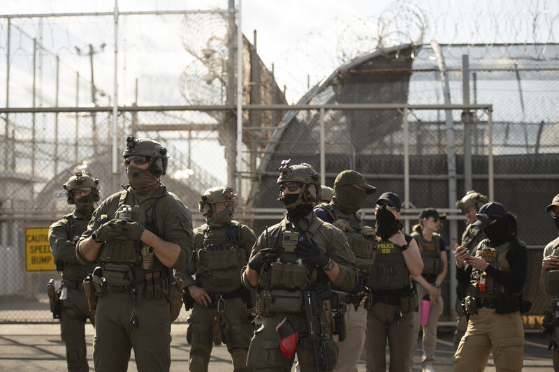 Tactical officers in tactical gear standing in formation at an industrial facility with chain-link fencing