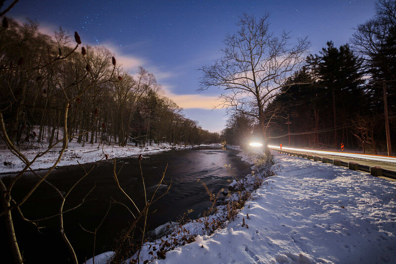 Snowy riverside landscape at dusk with bare trees lining both banks and car light trails on the right