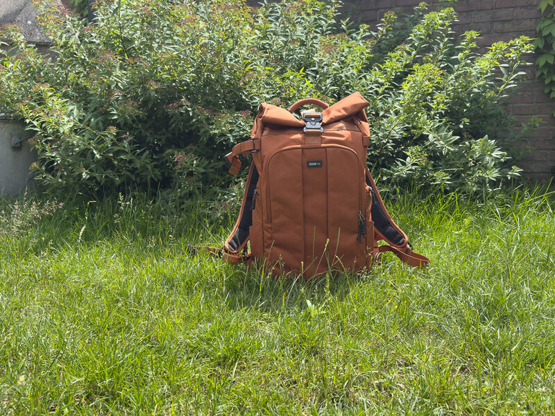 Rust-colored leather backpack sitting upright on grass with overgrown vegetation in background