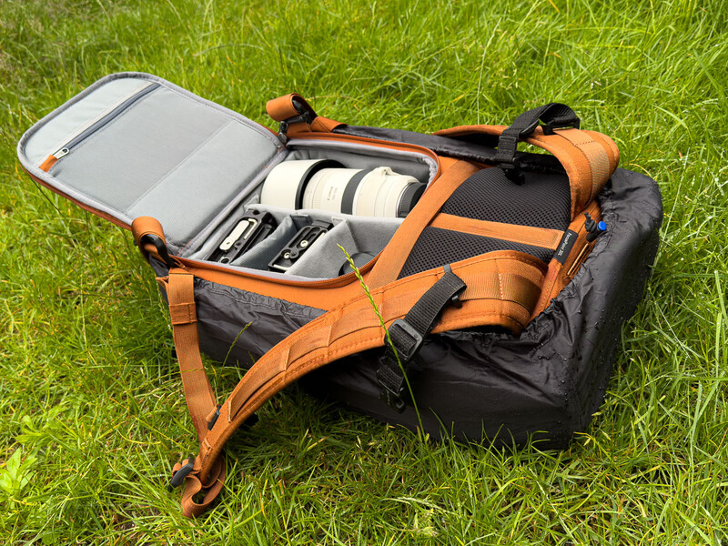 Orange and black camera backpack opened on grass displaying organized gear compartments with camera equipment inside