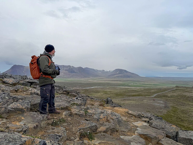 Hiker with orange backpack standing on rocky outcrop overlooking expansive tundra valley and distant mountains