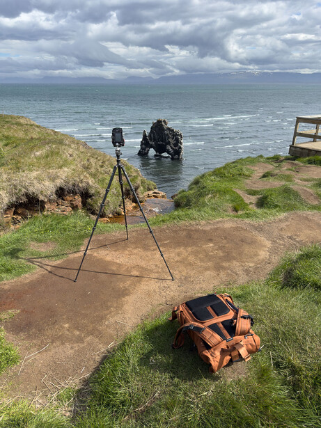 Camera on tripod positioned on coastal cliff overlooking sea with film crew visible in water below