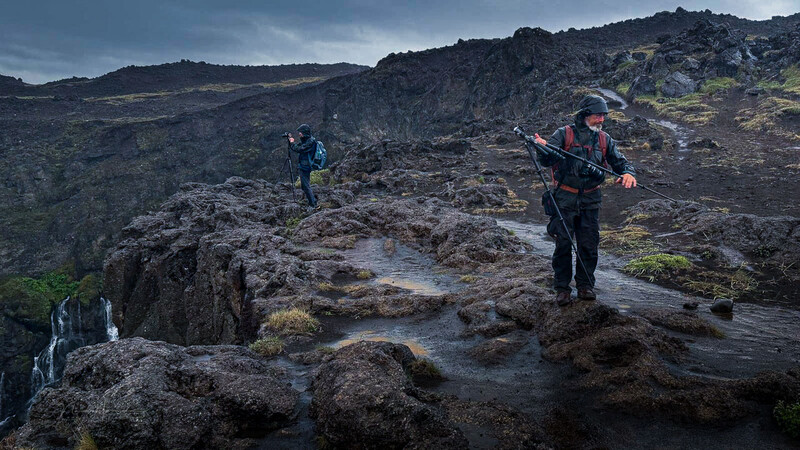 Three hikers traversing volcanic lava field with streams and moss-covered rocks
