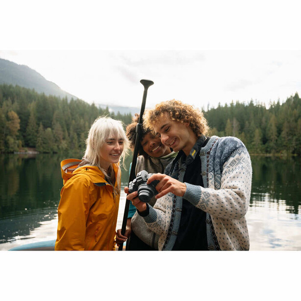 Three people examining a paddle by a calm alpine lake with forested mountains in background