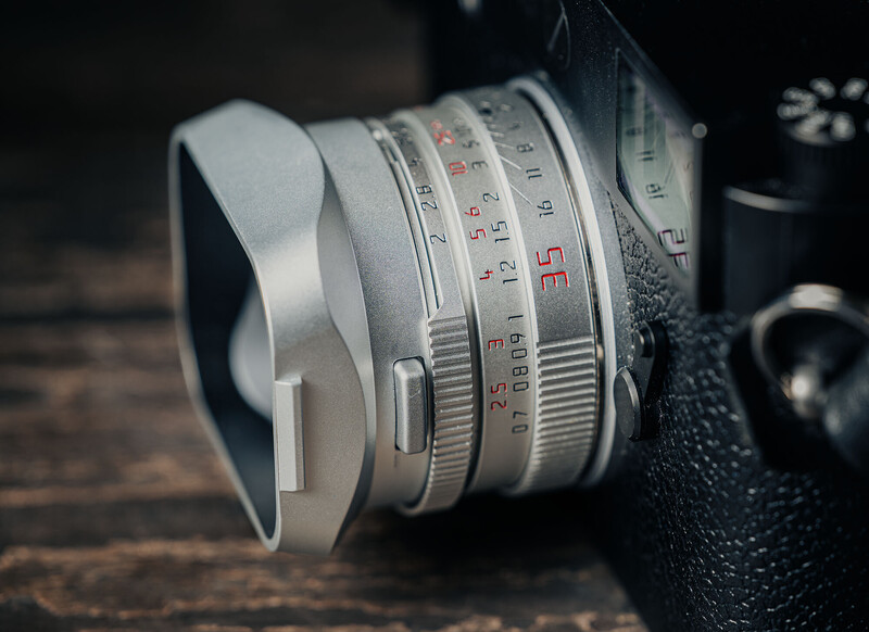 Silver vintage rangefinder lens with depth of field markings and focus ring, shown with lens hood attached