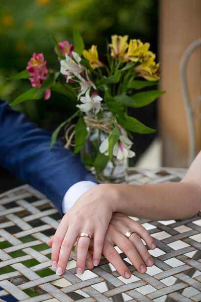 Bride and groom displaying wedding rings with hands resting on lattice table near colorful flower arrangement
