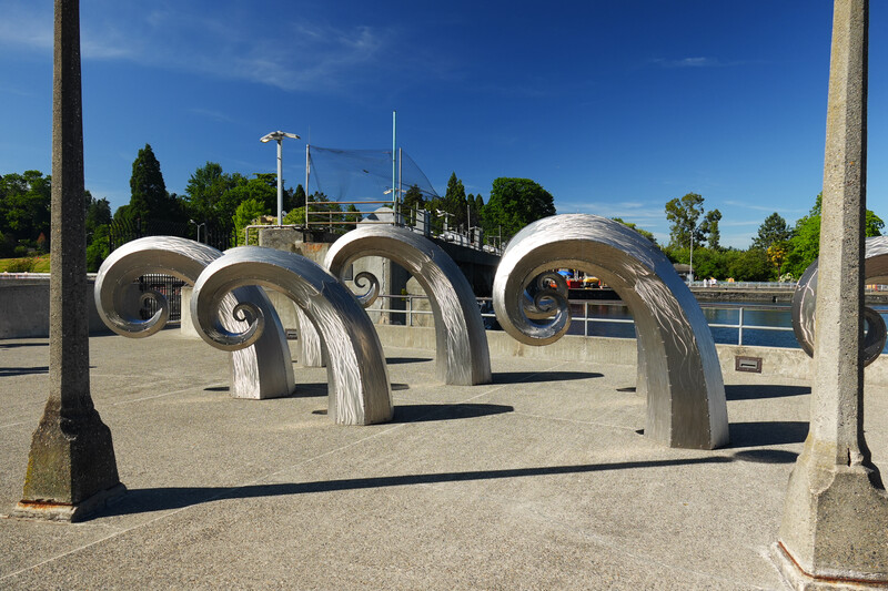 Three large metallic spiral arch sculptures in a concrete plaza with trees and water in background