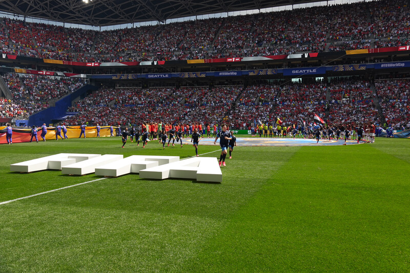 Large stadium filled with spectators during a soccer match, with players on the field and white geometric structures in foreground