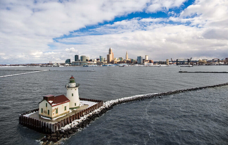 Red-roofed lighthouse on wooden pier extending into calm bay with city skyline in background