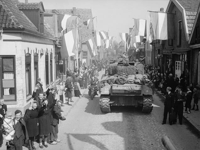 Military tanks advancing down a narrow European street lined with crowds of civilians