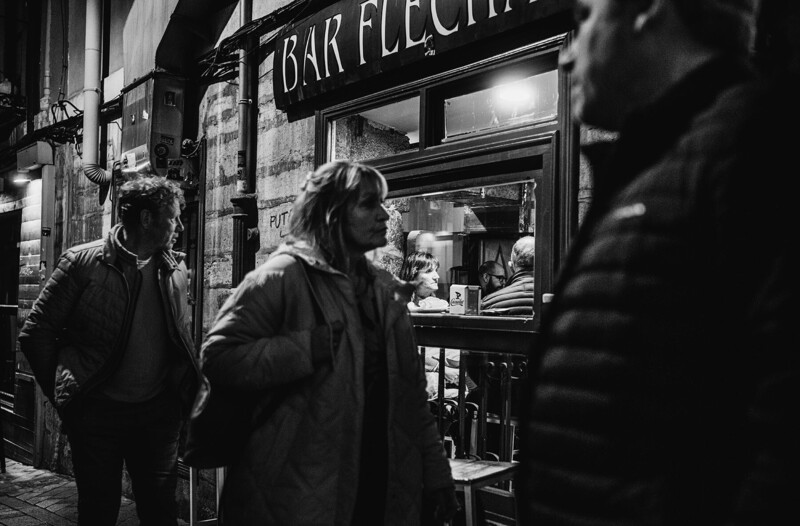 Nighttime street scene outside a bar with people in winter coats gathered near the entrance