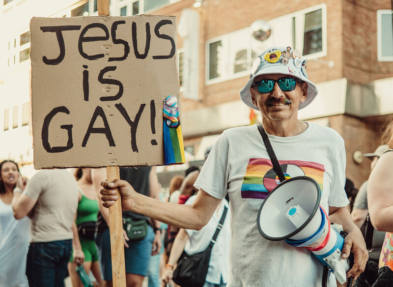 Man at outdoor pride parade holding megaphone and protest sign reading 'JESUS IS GAY!'