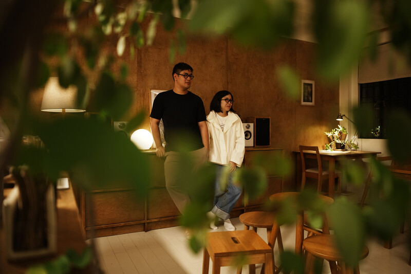 Two people standing in a warmly lit interior space with potted plants in foreground